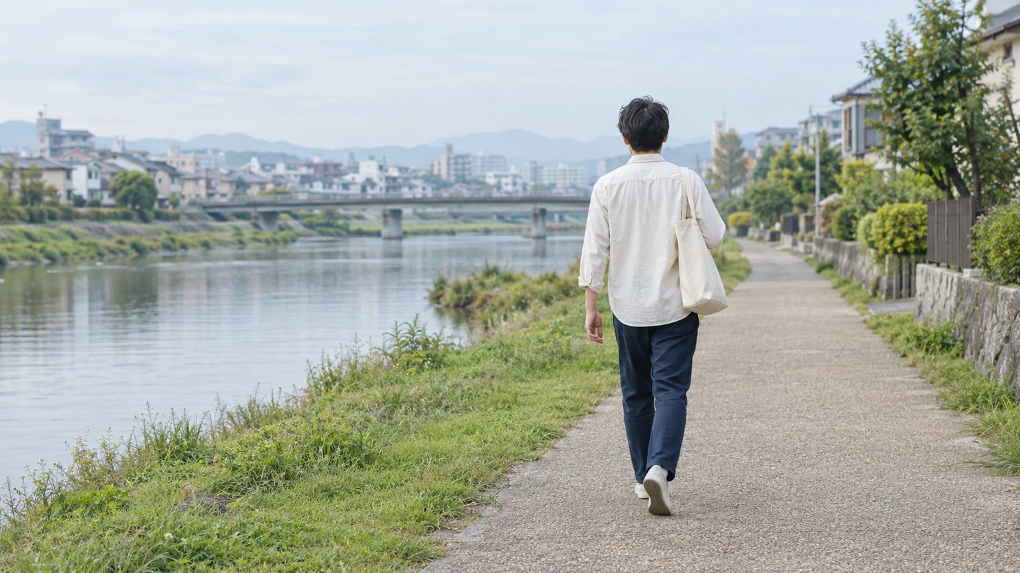 平日の朝、川沿いの遊歩道をトートバッグを提げて歩くエンジニアの後ろ姿