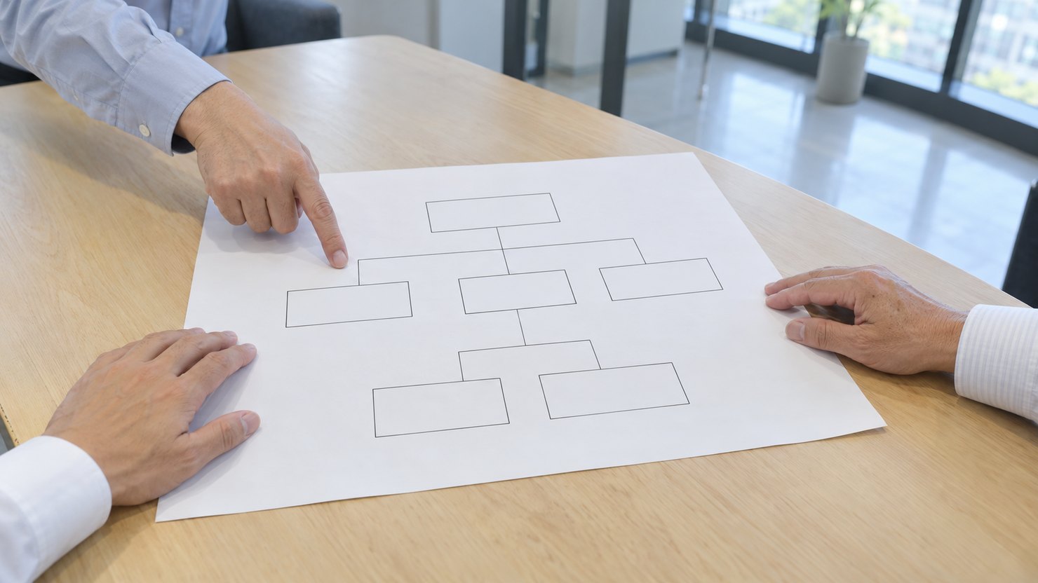 会議室の机で、四角と線だけの構成図の紙を3人の手が囲み、1人が指さしている