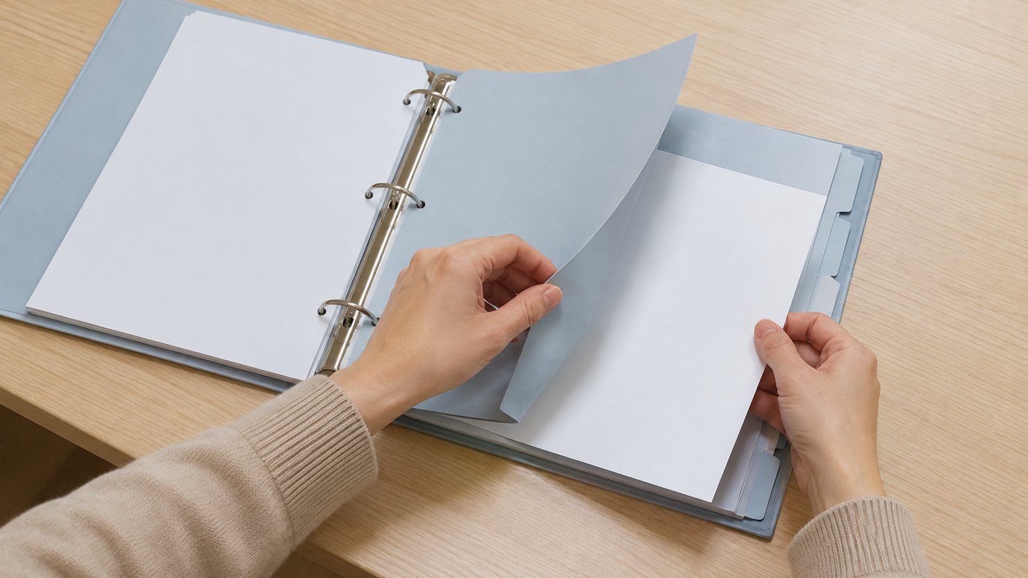 明るい机で、開いた無地のバインダーの仕切り紙の後ろに書類を差し込む両手