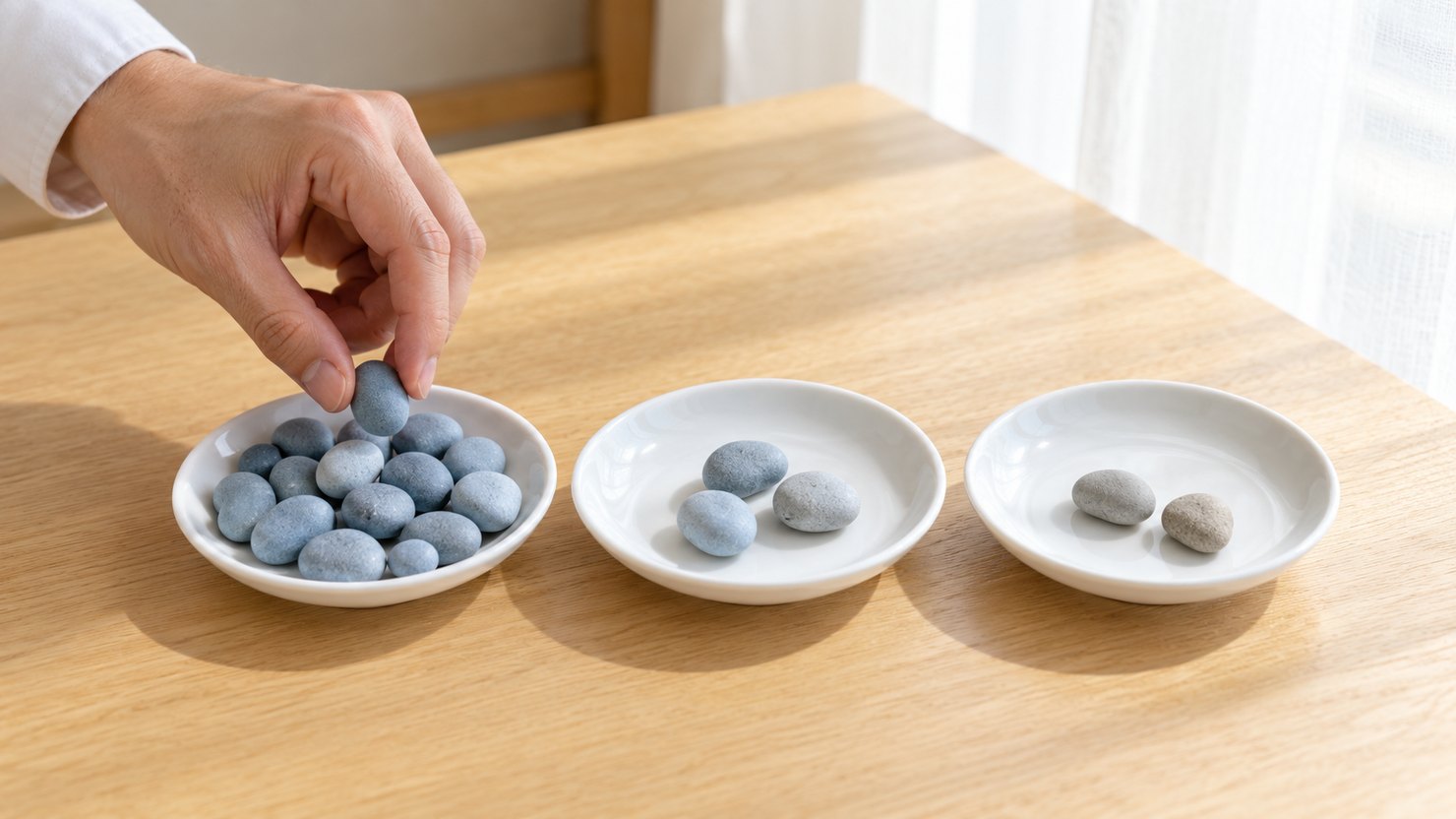 明るい机で、3つの白い小皿に小石を入れていき、左の皿だけが多くなっている手元