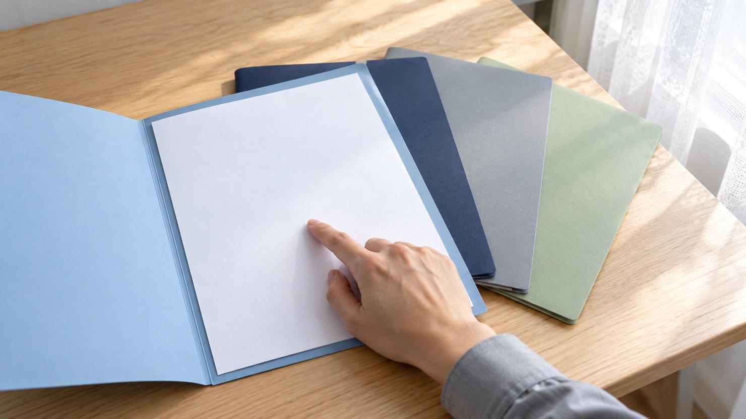 明るい窓際の机で、扇形に広げた4つの色違いの書類ばさみの1つを開き、中の紙を指でたどる手元