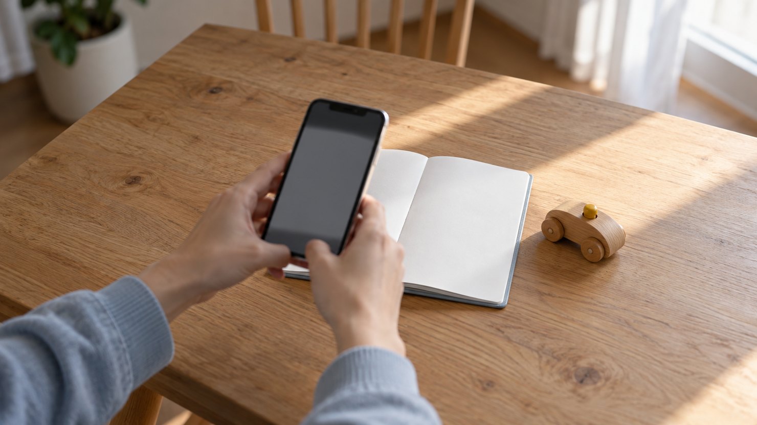 食卓で白紙の手帳をスマートフォンで撮影する両手と木のおもちゃの車